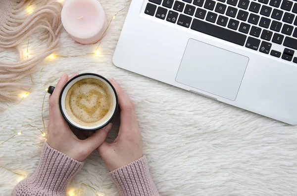Warm cozy winter workspace background. Flat lay. Girl holding a cup of coffee on fur fluffy white background with lights garland, candle and laptop. Top view mockup