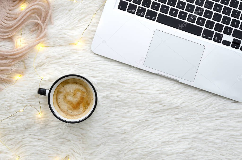 Warm cozy winter workspace with coffee and notebook on fur fluffy white background with lights garland and scarf. Top view mockup