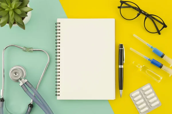 Medical or healthcare concept. Spiral blank notepad mock up on blue and yellow dual color background. Medical equipment, stethoscope. Flat lay view.