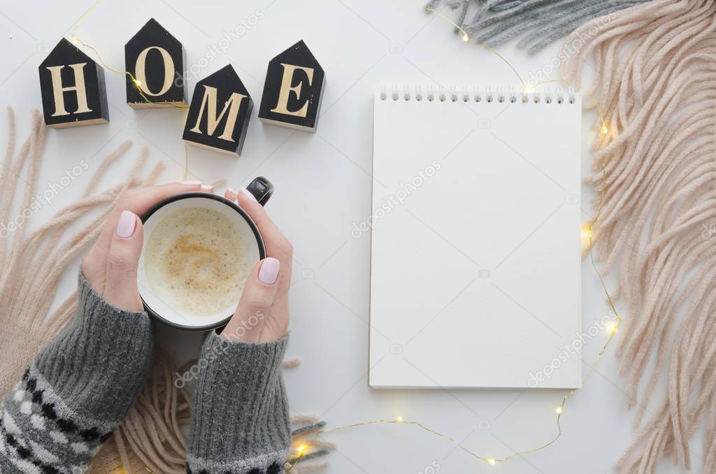 Womans hands with coffee and notepad mockup. Stylish home desk with scarf and lights. Cozy autumn or Winter concept. Flat lay, top view