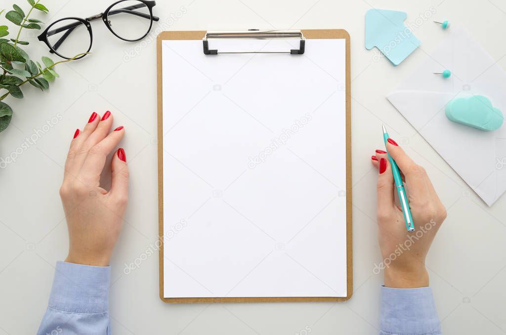 Business woman in blue shirt holds pen. lipboard mockup A4 size isolated on white desk, office supplies, eyeglasses, envelope. Top view flat lay. Template for reports, resume, brief, form, contract