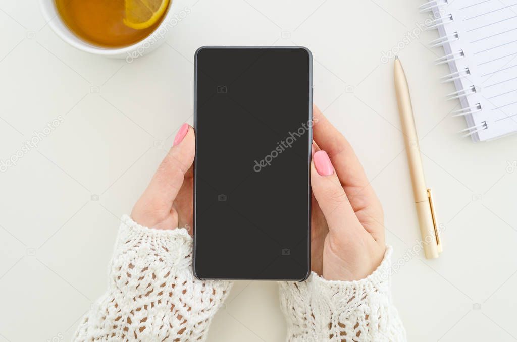 Mock up smartphone Top view. Student woman holds with two hands smartphone. Workplace, mobile phone, tea, pen and notepad