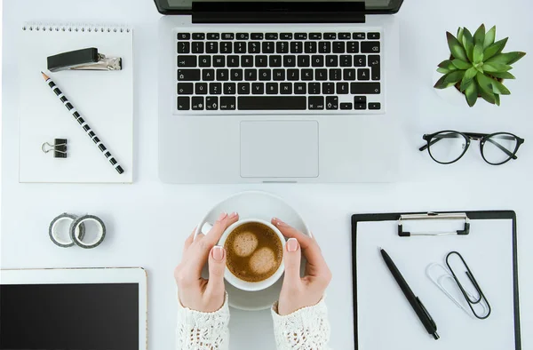 Top view office desk. Workspace with girls hands, laptop, notepad, clipboard, tablet, coffee cup, eye glasses,pen and clips. Flat lay. Desktop concept.