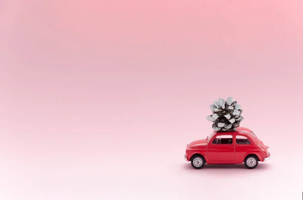 Red Retro toy model car with christmas pine cone on pink background. christmas, winter delivery concept