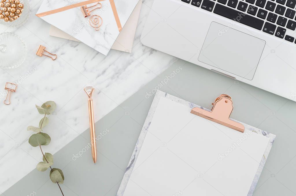 Office desk table with laptop, clipboard, rose gold pen, clips and supplies, pretty eucalyptus branches on marble gray background. Top view with copy space