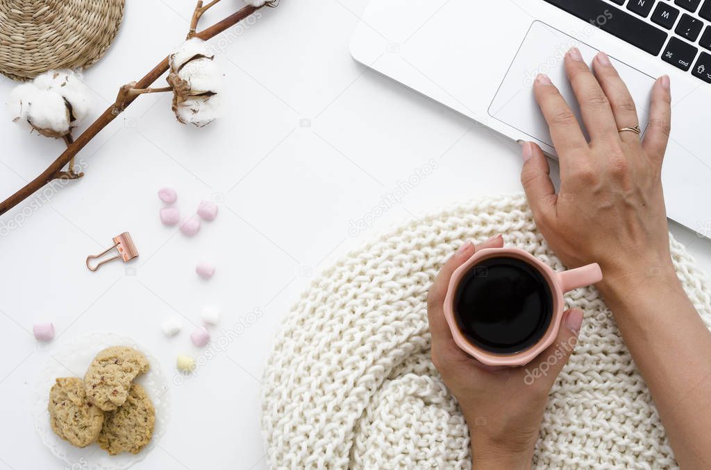 A woman is working behind a laptop and is holding coffee. On the table lie oatmeal cookies, marshmallow and cotton flowers. Autumn or Winter concept. Flat lay, top view