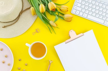 Womens sarı arka plan bloglama. Çay bardağı, pano, bahar lale buket, şapka ve sarı arka planda klavye ile ofis masası çalışma. Kopya alanı ile üst görünüm