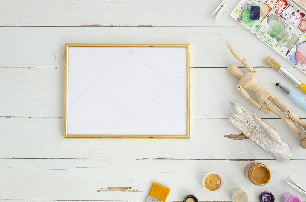 Creative artist workplace mockup. Top view on table with blank sheet of paperin golden frame with paintbrushes. Copy space for text on white wooden planks background