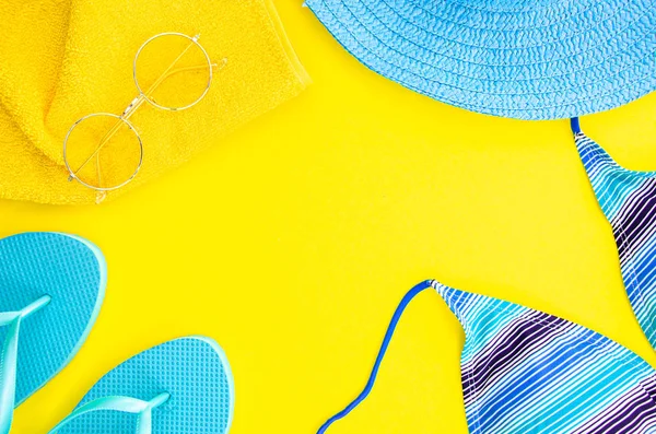 Summer flat lay mockup. Blue flip flops, palm leaf and cloth with sunglasses on yellow background. Womens blue hat and bikini. Top view frame with copy space for text or lettering