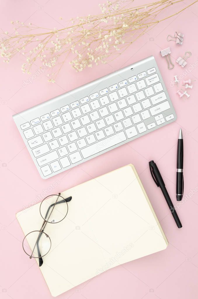 Overhead view desktop with computer keyboard, reading glasses, opened blank notepad and pens. Flat lay mockup still life concept on pink background