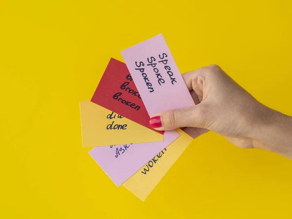 Mão feminina segura flashcards com verbos ingleses irregulares escritos à mão isolados em amarelo. Conceito de aprendizagem de línguas — Fotografia de Stock