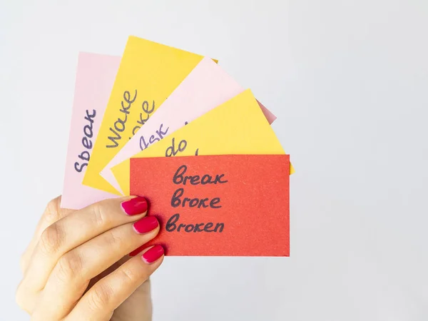 Vrouwelijke hand houdt flashcards met handgeschreven onregelmatige Engelse werkwoorden geïsoleerd op grijs. Leerconcept voor vreemde talen — Stockfoto