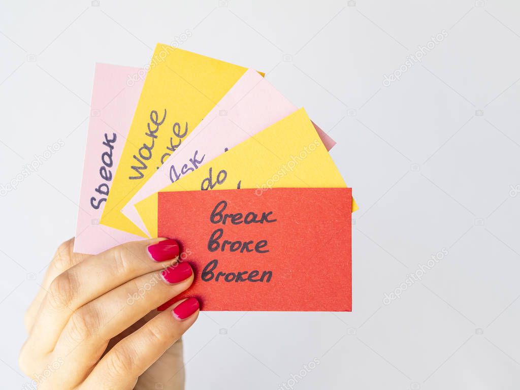 Female hand holds flashcards with handwritten irregular English verbs isolated on grey. Foreign language learning concept