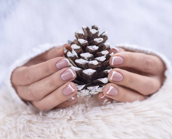 Winter SPA and manicure concept. Beautiful female hands in white knitted sweater with manicure holds pine cone — Stock Photo, Image