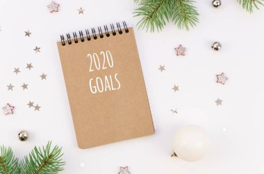 Düz Yeni Yıl hedefleri kavramı. Üst manzara sarmal not defteri ve 2020 gol işareti. Gelecek yıl için köknar ağacı dalları ve beyaz masa üzerindeki xmas süslemeleri içeren defter yapmak için