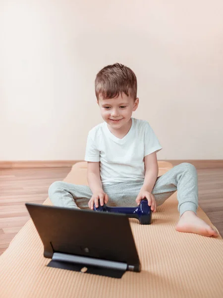4 yaşında bir çocuk, halterli spor egzersizleri yapmak için tablet bilgisayar kullanıyor. Evde jimnastik — Stok fotoğraf