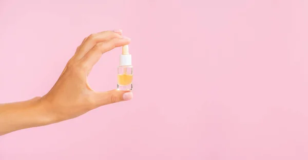 beauty products banner with copy space. Dropper bottle with oil mockup. Female hand holds yellow cosmetic oil in a clear jar on a pink background