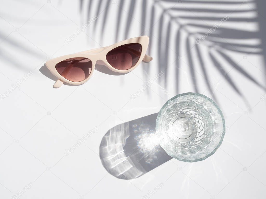 Summer minimal concept. Top view clear glass and fashion sunglasses with soft shadows of tropical palm leaves on a white background