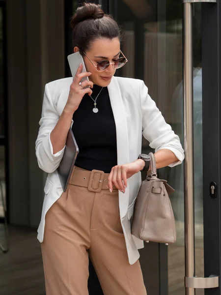 Mujer ocupada en traje blanco mirando su reloj y hablando por teléfono mientras sale del edificio de negocios — Foto de Stock