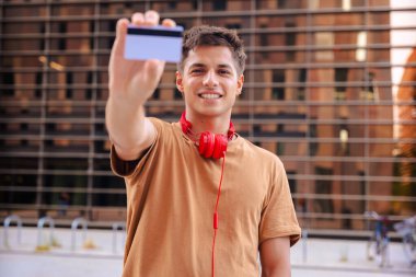 Boynunda kırmızı kulaklıklarla gülümseyen genç bir adam açık havada kameraya boş bir kartvizit gösteriyor. Metal parmaklıklar ve arkadaki bulanık binalarla modern bir şehir arka planının önünde duruyor. 