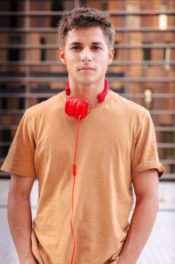 Boynuna kırmızı kulaklıklar takmış, modern mimari ve güneş ışığını yansıtan şehir ortamında duran kameraya içten bir şekilde gülümseyen genç bir adamın portresi.