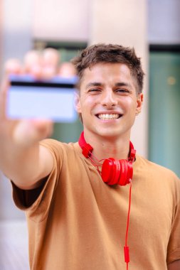 Boynunda kırmızı kulaklıklarla gülümseyen genç bir adam açık havada kameraya boş bir kartvizit gösteriyor. Metal parmaklıklar ve arkadaki bulanık binalarla modern bir şehir arka planının önünde duruyor. 