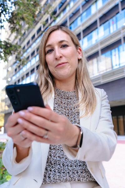 Confident business woman outdoors in white suit using smartphone, modern glass building, urban city, low angle, career, professional, technology, digital communication, working, leadership, success 