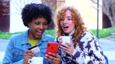 İki çok ırklı bayan arkadaş açık kafede oturuyor, akıllı telefon ve kredi kartını online alışveriş için kullanıyor, kahve içiyor, gülümsüyor, eğleniyor, dijital ödeme yapıyor, teknoloji, arkadaşlık, modern yaşam tarzı.