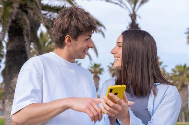 Palmiye ağaçlarının altında deniz kenarında oturan genç çift, birlikte sarı bir akıllı telefona bakarken gülümsüyor, ekrana işaret eden erkek arkadaş, aşkı, seyahat anlarını ve bugün dijital bağlanmayı yakalıyor. 