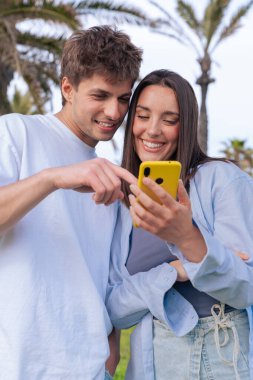 Palmiye ağaçlarının altında deniz kenarında oturan genç çift, birlikte sarı bir akıllı telefona bakarken gülümsüyor, ekrana işaret eden erkek arkadaş, aşkı, seyahat anlarını ve bugün dijital bağlanmayı yakalıyor. 