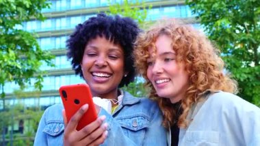 Kentsel parkta gülümseyen ve akıllı telefona bakan iki genç kadın, yaşam tarzı teknolojisi, çevrimiçi iletişim ve sosyal medya