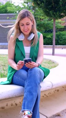  Yeşil yelekli genç sarışın kadın eğri büğrü park bankında boynunda kulaklıklarla oturuyor. Açık havada dinlenirken akıllı telefonuna gülümsüyor. Modern dijital yaşam tarzını ve kentsel rahatlığını yansıtıyor.