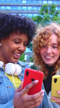 Açık havada akıllı telefonları dikey video formatında kullanırken gülümseyen çok ırklı genç kadınlar, yaşam tarzı, arkadaşlık, teknoloji, sosyal bağlantı, eğlence, iletişim