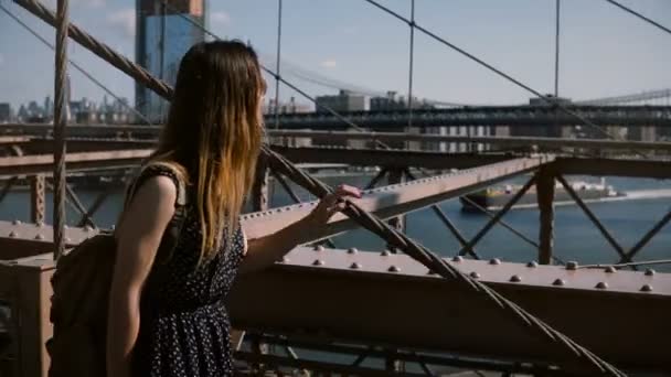 Camera follows young happy female tourist with backpack in sunglasses walking along Brooklyn Bridge, enjoying view 4K. — Stock Video