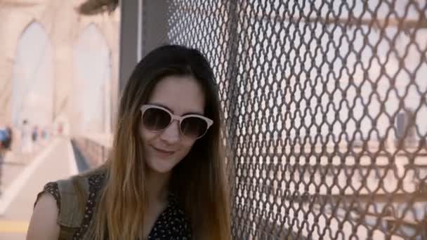 Belle touriste féminine en lunettes de soleil souriant, penchée vers la clôture du filet d'ombre au pont de Brooklyn, marchant vers la caméra 4K . — Video