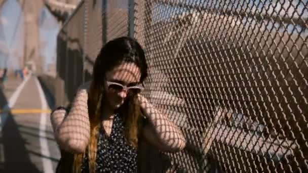 Schöne europäische Frau mit stylischer Sonnenbrille, die vor die Kamera läuft, Haare am Brooklyn Bridge Schattenzaun berührt 4k. — Stockvideo