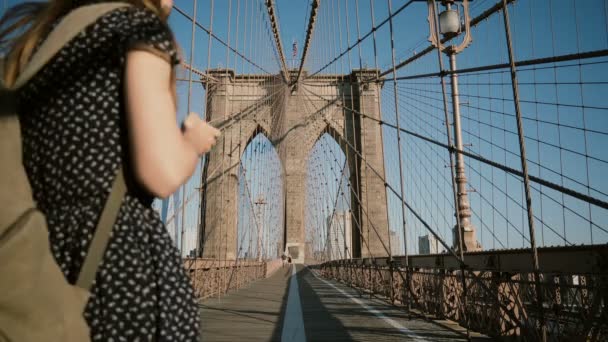 Young European female tourist with backpack walking along Brooklyn Bridge, New York and looking at smartphone 4K. — Stock Video
