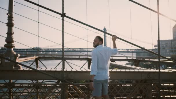 Adulto caucasiano homem de pé em Brooklyn Bridge à espera de alguém com os braços dobrados, olhando para pulseira de relógio inteligente 4K — Vídeo de Stock