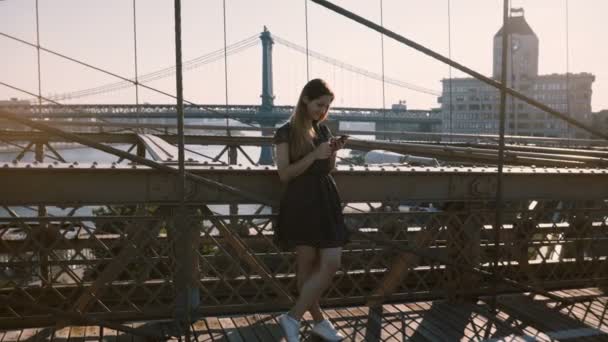 Piękna kobieta blogger Kaukaska, za pomocą aplikacji na smartfony w Brooklyn Bridge nad rzeką panorama, a następnie odchodzisz 4k. — Wideo stockowe