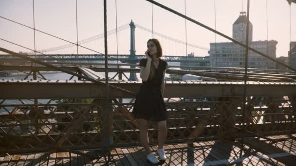 Feliz chica caucásica hablando por teléfono, de pie cerca del increíble puente de Brooklyn, Nueva York vista en un hermoso día de verano 4K — Vídeo de stock
