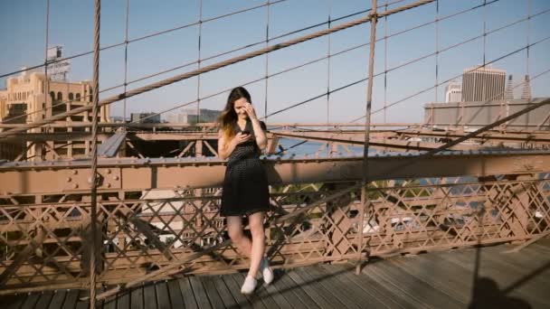 Hermosa blogger europea está usando la aplicación de compras de teléfonos inteligentes en Brooklyn Bridge, Nueva York en un día soleado 4K . — Vídeos de Stock