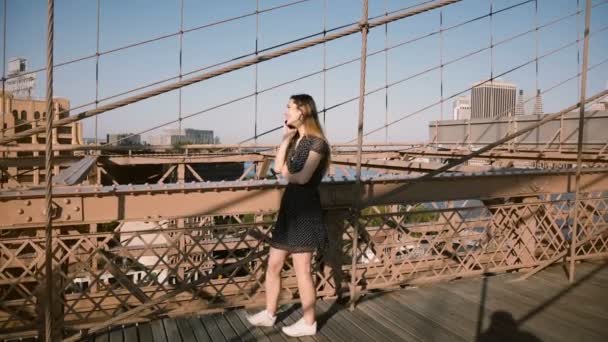 Bastante sonriente chica coqueta caucásica hablando con un amigo por teléfono en el puente de Brooklyn, caminando lentamente hacia adelante y hacia atrás 4K — Vídeo de stock
