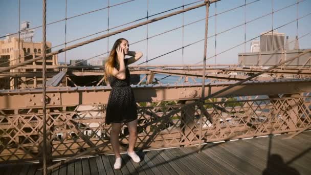 Szczęśliwy Europejskiej młoda kobieta za pomocą smartfona, a następnie wykonanie rozmowy telefonicznej i rozmowy z przyjacielem na Brooklyn Bridge, Nyc 4k. — Wideo stockowe