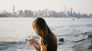 Tanınmayan kadın uzun saçlı günbatımı 4 k New York şehir silüeti manzaralı nehir plajda smartphone app kullanarak.