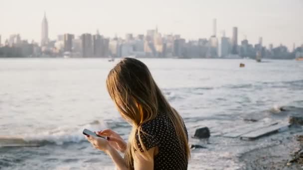 Femme méconnaissable aux cheveux longs utilisant une application smartphone à la plage de la rivière avec vue sur les toits de la ville de New York au coucher du soleil 4K .