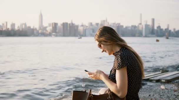 Aantrekkelijk meisje van de Kaukasische in zonnebril met smartphone messenger app, kijken rond New York rivier strand 4k. — Stockvideo