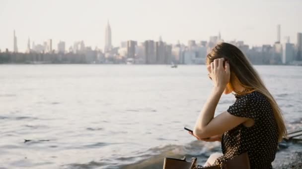 Belle fille européenne avec les cheveux longs dans les lunettes de soleil en utilisant smartphone shopping application en ligne à New York coucher de soleil plage 4K .