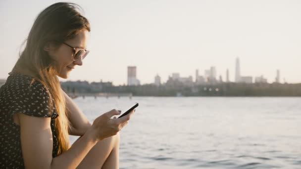 Gondolkodó kaukázusi lány napszemüveg használ smartphone társadalmi hálózat app, élvezi a csodálatos naplementét a városi strand 4k. — Stock videók