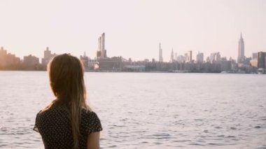 Güneş gözlüğü güzel kız saçları rüzgarda tek başına duruyorsa, nehir kıyısına New York günbatımı 4 k yürüyor..