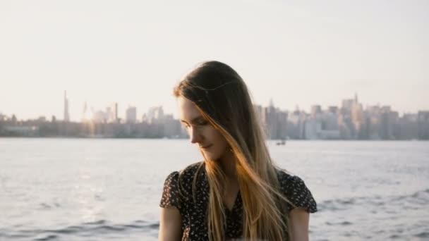 Ritratto di felice ragazza europea con capelli volanti in posa, sorridente alla macchina fotografica sulla spiaggia al tramonto con skyline di New York City 4K — Video Stock